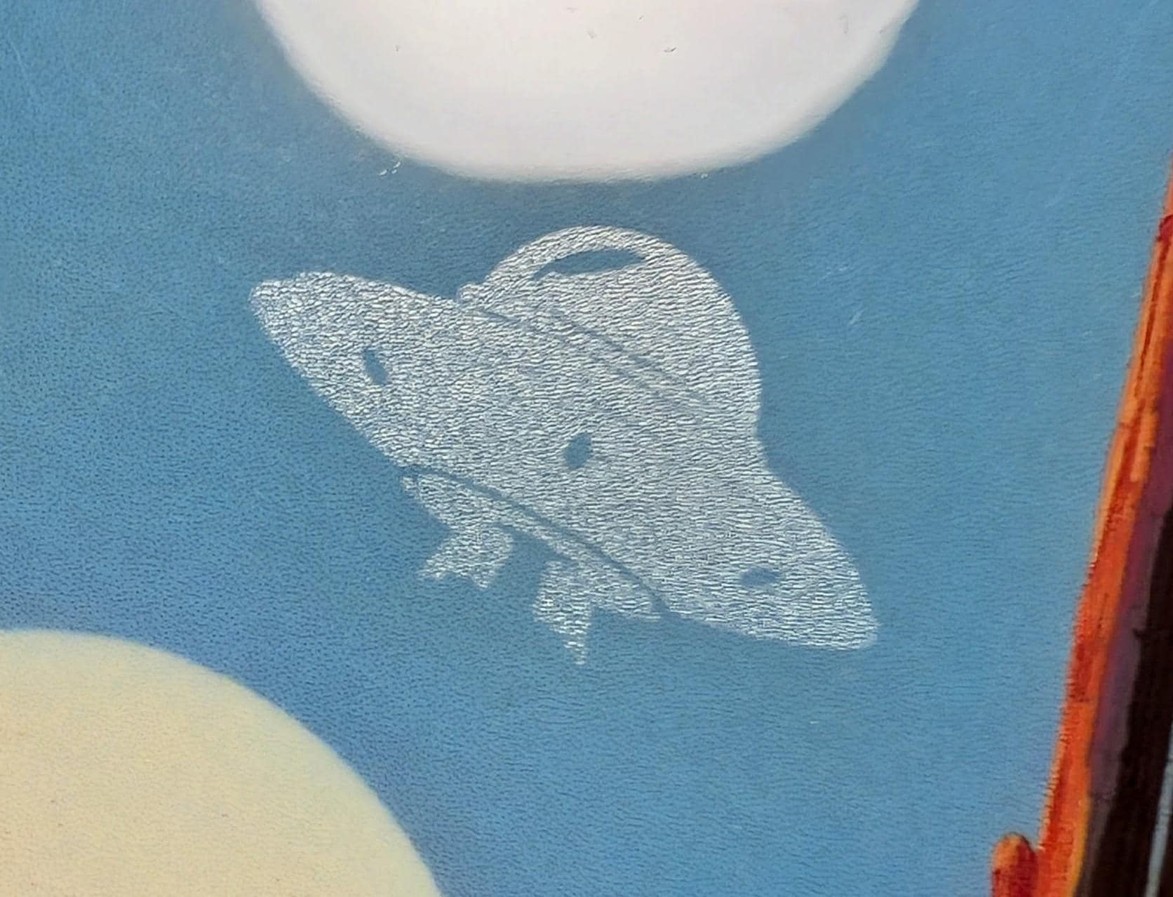 Extreme close-up of a custom phone case. A retro UFO is precision laser-engraved into the blue sky. The detail shot highlights the strong contrast between the smooth, colorful sublimation print and the fine, tactile texture of the laser engraving.