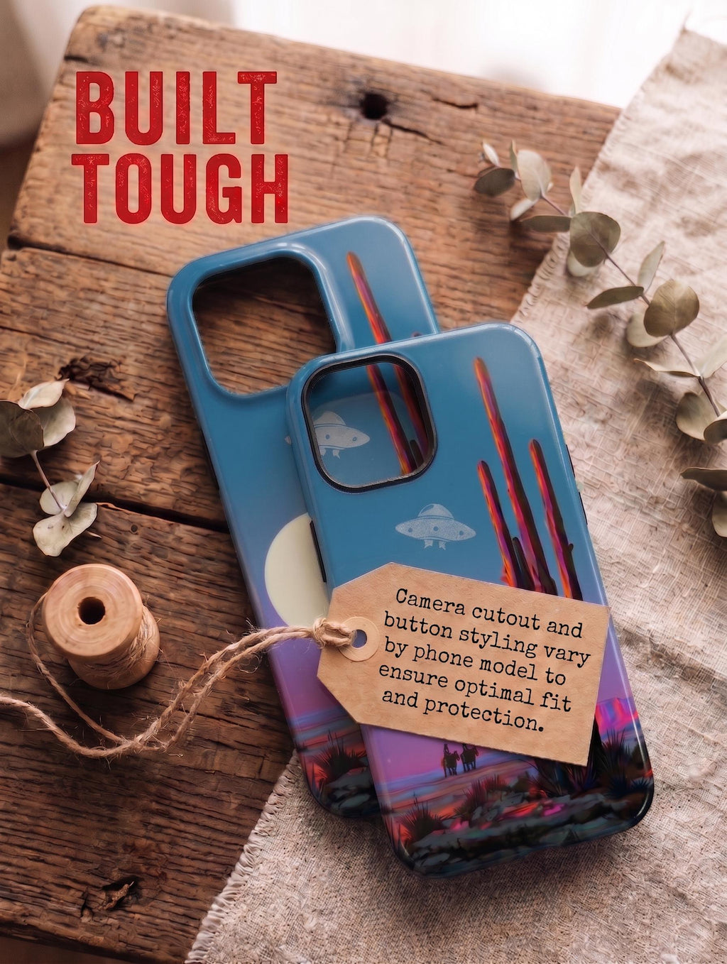 Top-down flatlay of two stacked smartphone cases on a rustic wooden table. Both cases feature a desert landscape design with a saguaro cactus, UFO, cowboys, and a sunset moon. Red bold text at the top left reads 'VARIANTEN'. The top case has a brown kraft paper tag tied with jute twine, containing German text explaining that camera cutouts and button designs vary by phone model for optimal fit and protection. A wooden thread spool and dried eucalyptus branches rest on linen fabric nearby