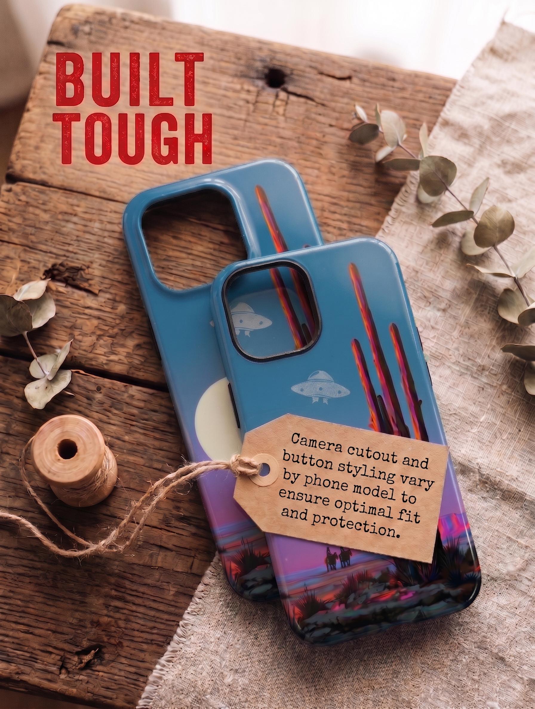 Top-down flatlay of two stacked smartphone cases on a rustic wooden table. Both cases feature a desert landscape design with a saguaro cactus, UFO, cowboys, and a sunset moon. Red bold text at the top left reads 'VARIANTEN'. The top case has a brown kraft paper tag tied with jute twine, containing German text explaining that camera cutouts and button designs vary by phone model for optimal fit and protection. A wooden thread spool and dried eucalyptus branches rest on linen fabric nearby